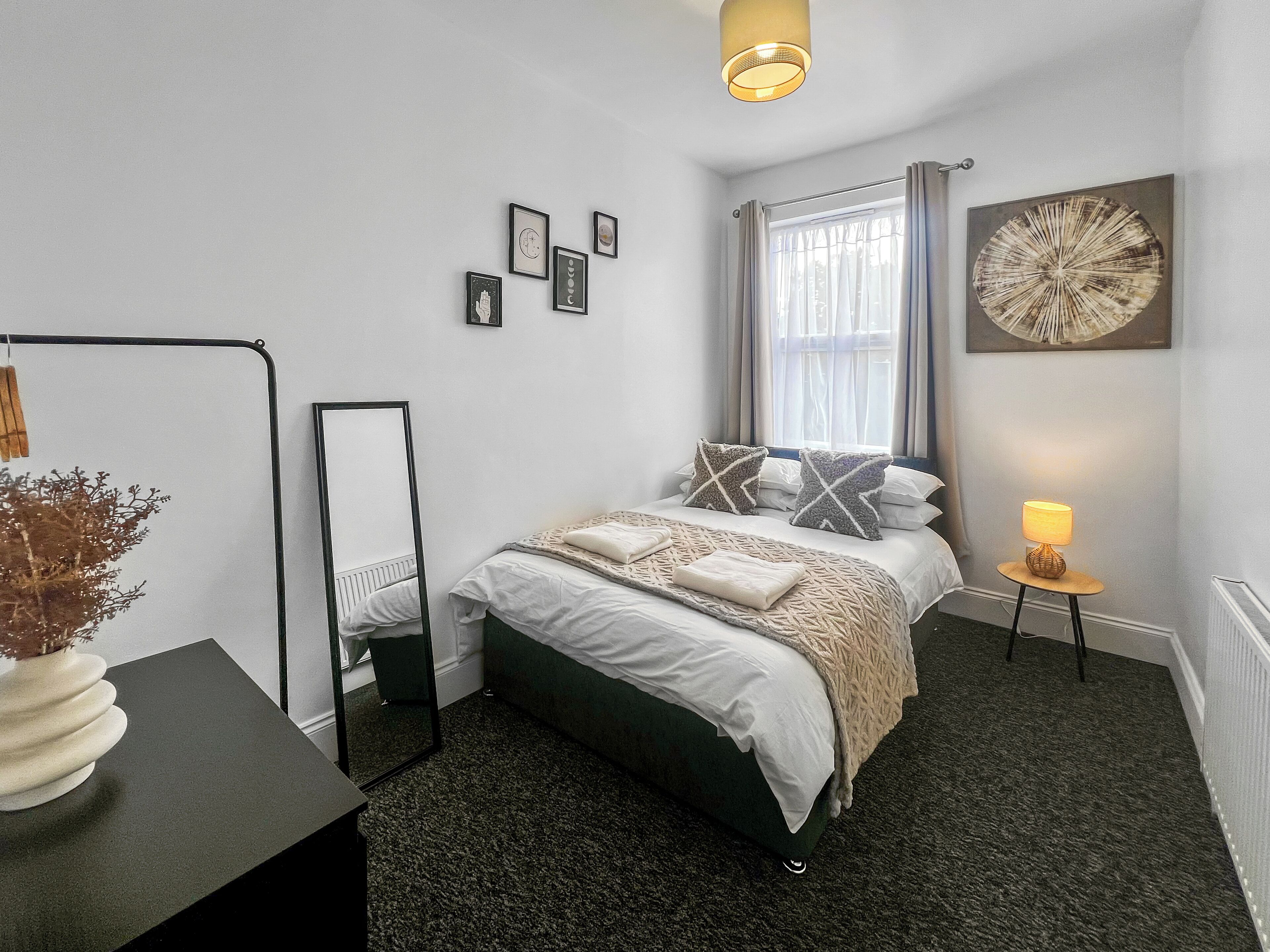 A neatly arranged bedroom with a bed, a bedside table, a lamp, a mirror, and wall art.