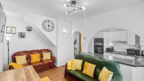 Una sala de estar moderna con un sofá de cuero marrón y un sofá verde con almohadones amarillos, una mesa de comedor y una cocina con armarios blancos y horno empotrado.