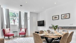 Un espace repas moderne avec une table ronde pour quatre personnes, un téléviseur à écran plat fixé au mur et une fenêtre avec des rideaux.