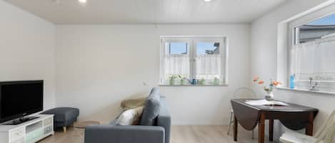 A modern living room with a flat-screen TV, grey sofa, wooden dining table, and a window with curtains.