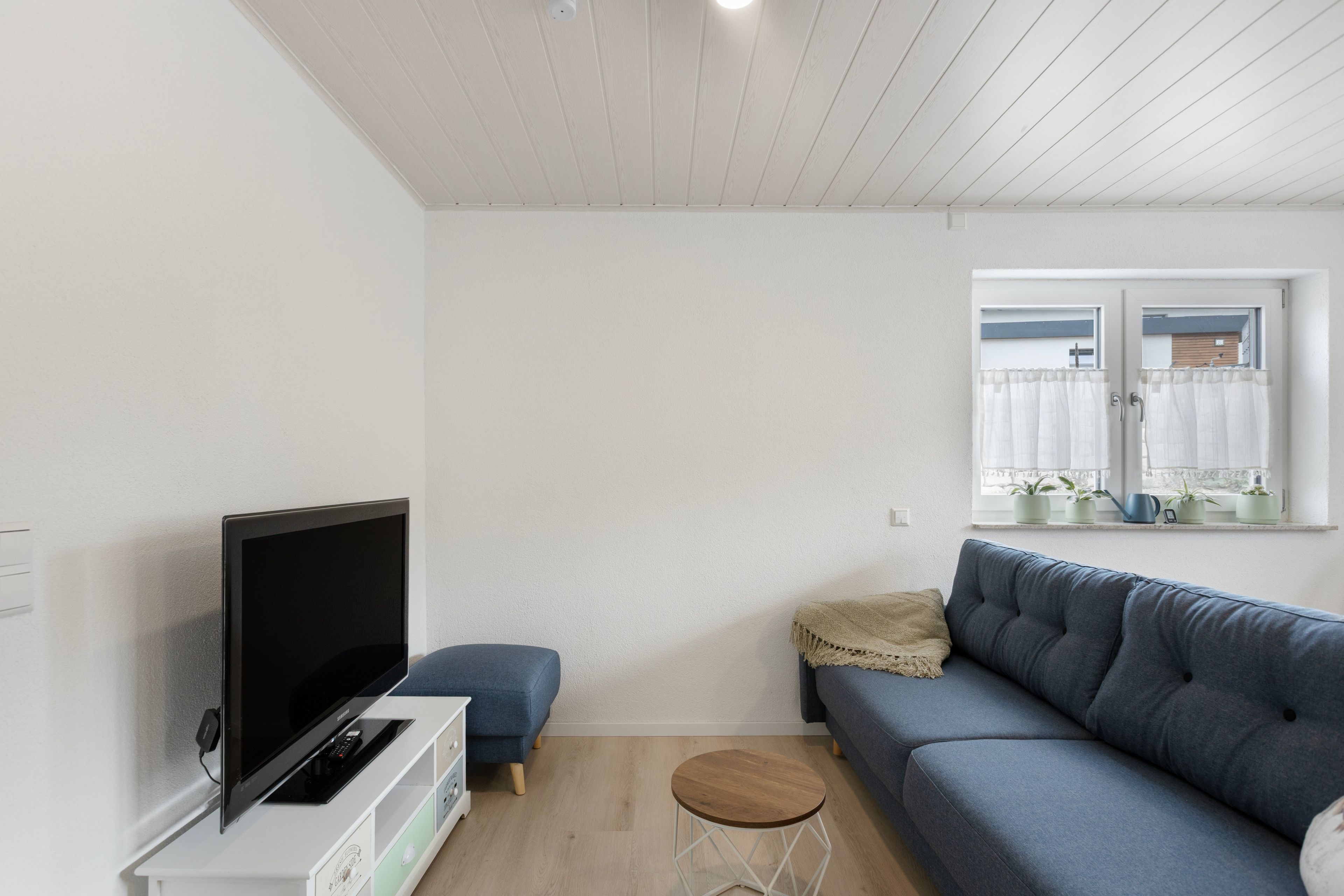 Ein modernes Wohnzimmer mit einem Flachbildfernseher, einem blauen Sofa, einem kleinen Holzschemel und einem Fenster mit Pflanzen.