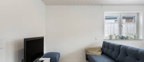 A modern living room with a flat-screen TV, a blue sofa, a small wooden stool, and a window with plants.