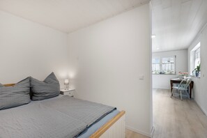 Une chambre à coucher comprenant un lit, une table de chevet, une lampe et donnant sur la pièce adjacente.