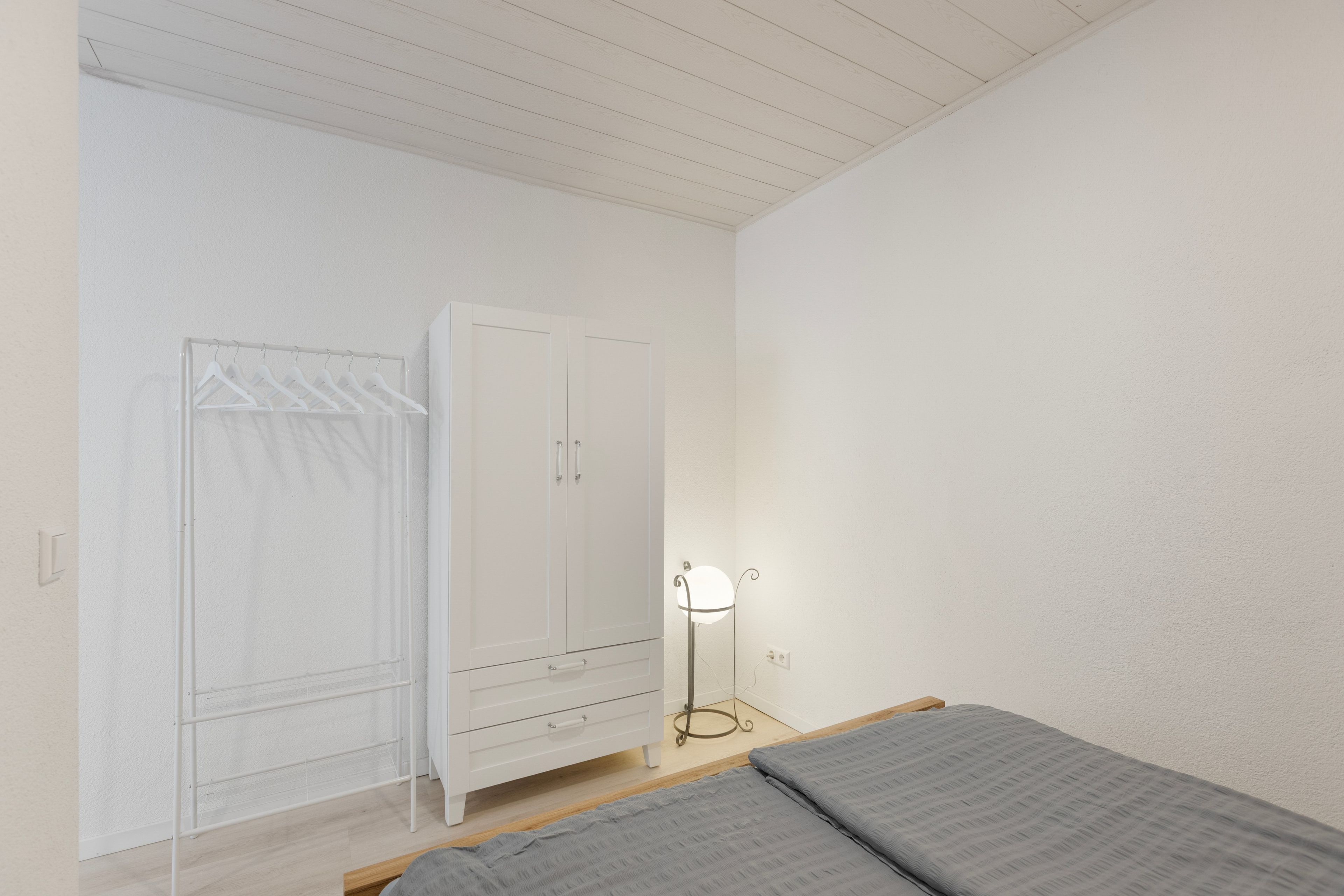 A bedroom with a bed, a wardrobe, a hanging clothes rack, and a floor lamp.