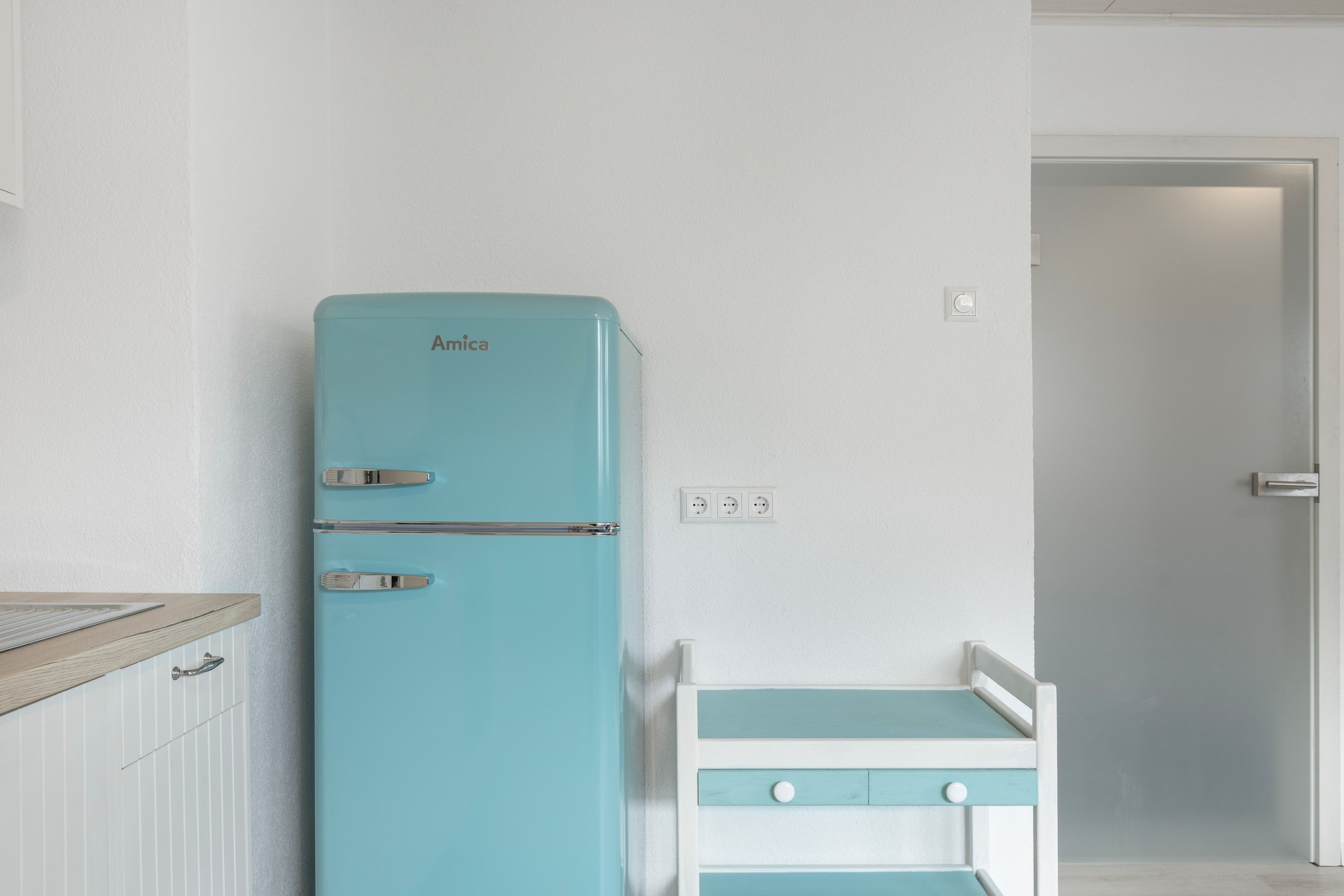 Eine kleine Küchenzeile mit einem türkisblauen Amica-Kühlschrank, einer weißen Arbeitsplatte und einer Tür zu einem anderen Raum.