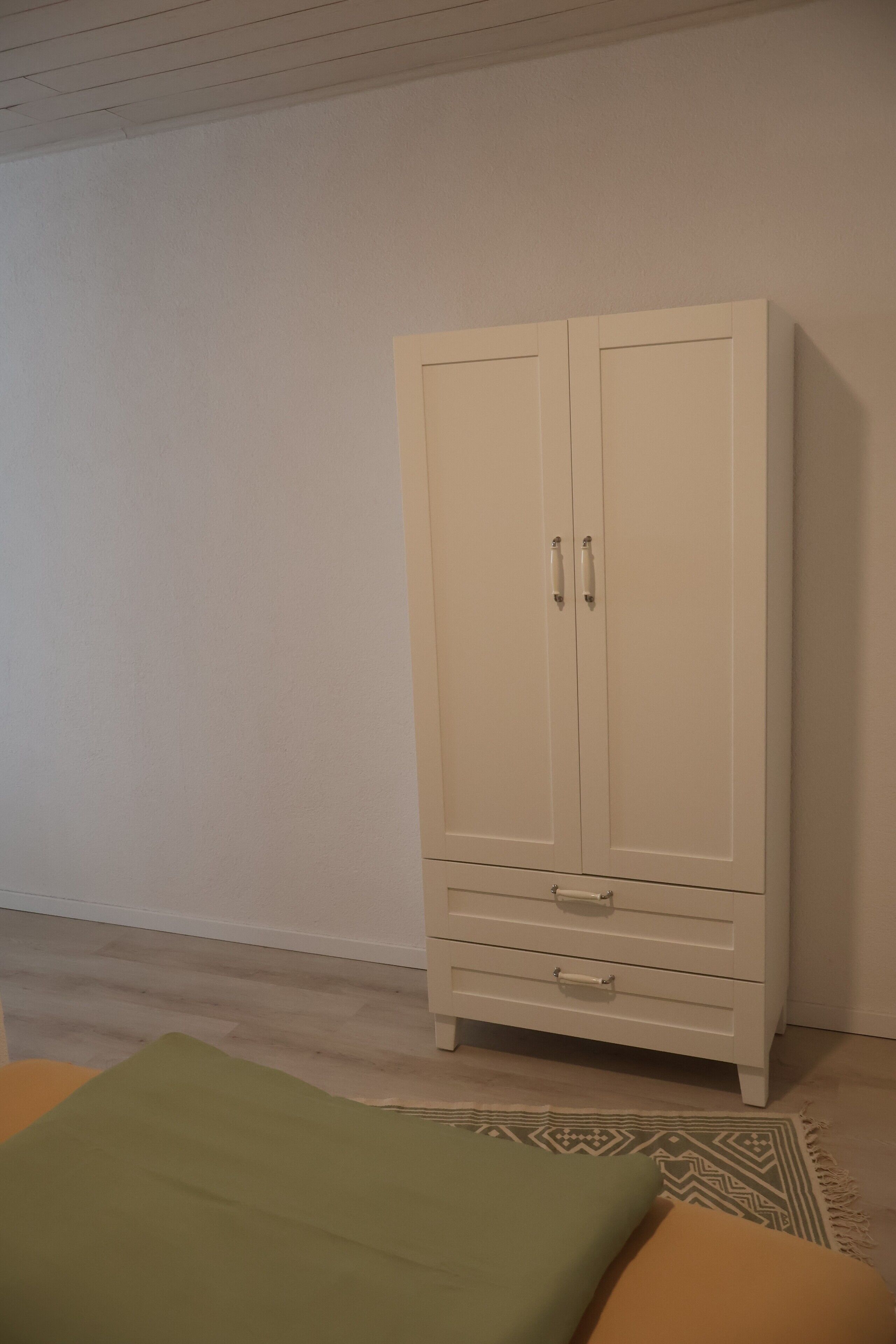 Une armoire blanche à deux portes et un tiroir, un tapis à motifs et un coussin vert et jaune sur un plancher en bois.