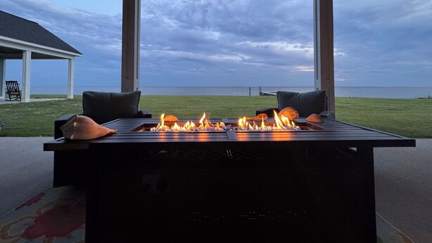 Ein Zimmer mit großem Fenster mit Blick auf eine Wiese und ein Gewässer. In der Mitte des Raumes befindet sich eine Feuerstelle.