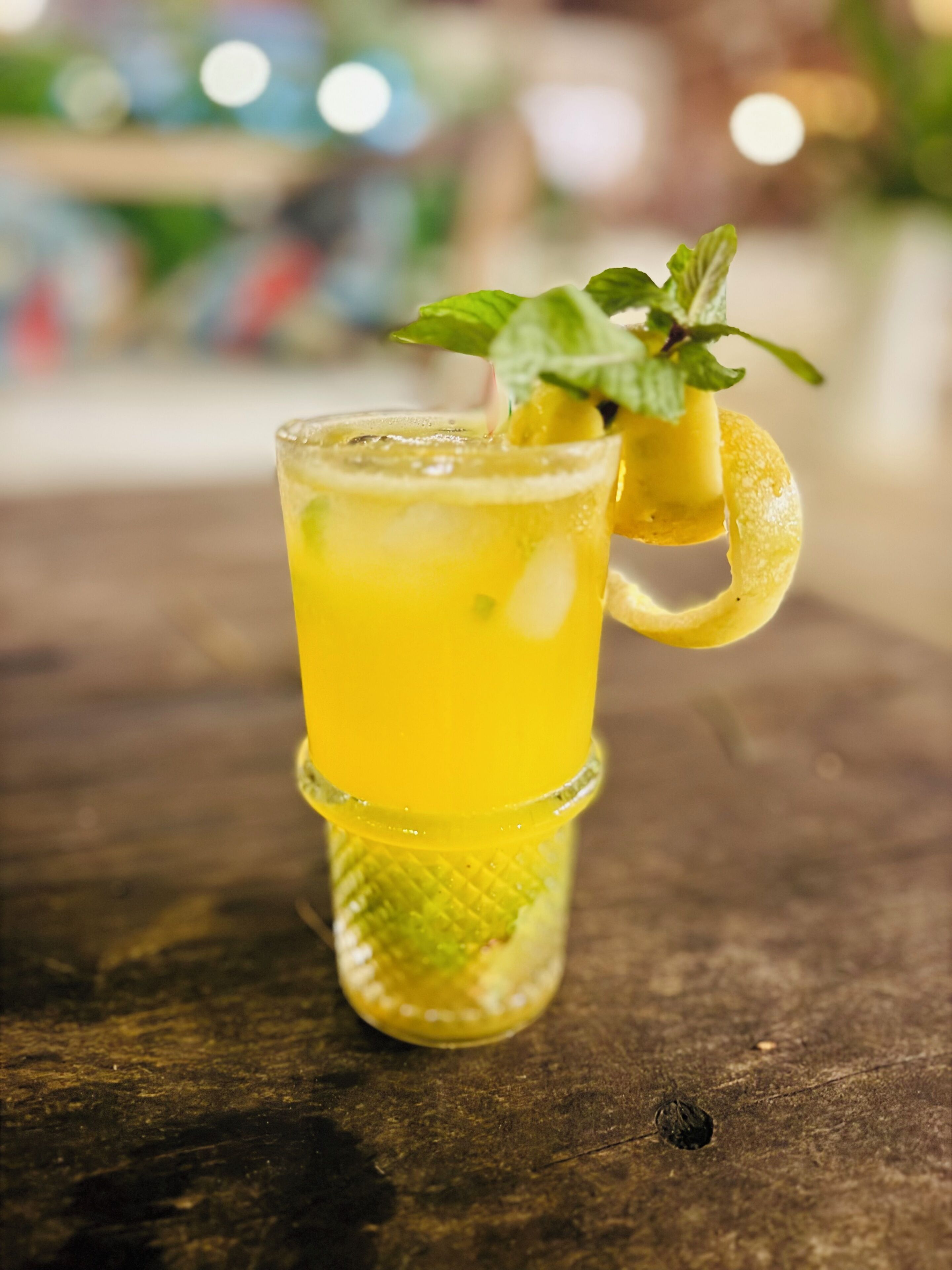 Un vaso de bebida amarilla con una rodaja de limón y una ramita de menta sobre una mesa de madera.