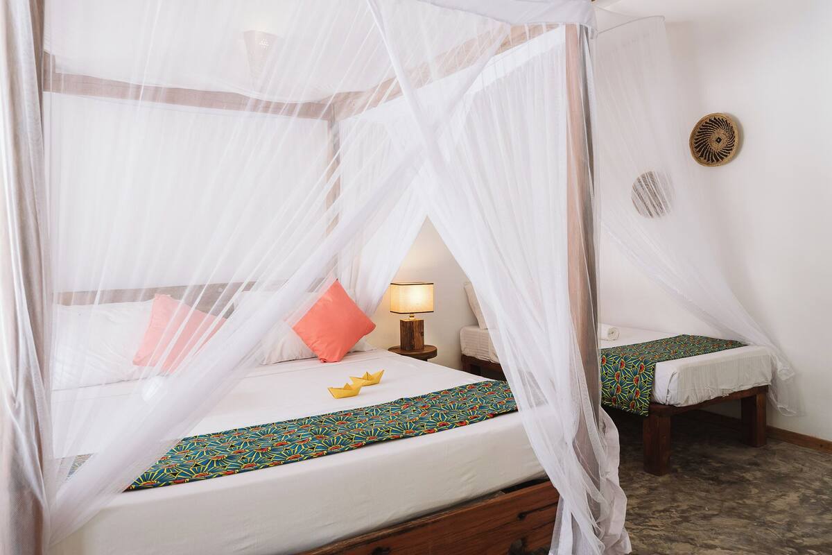 A room with a bed under a mosquito net, a bedside table with a lamp, and a bench with a patterned bedspread.