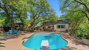 A swimming pool with a wooden deck, surrounded by trees and a house.