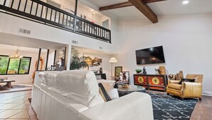 A spacious living area with a high ceiling, a balcony, a flat-screen TV, and a leather armchair.