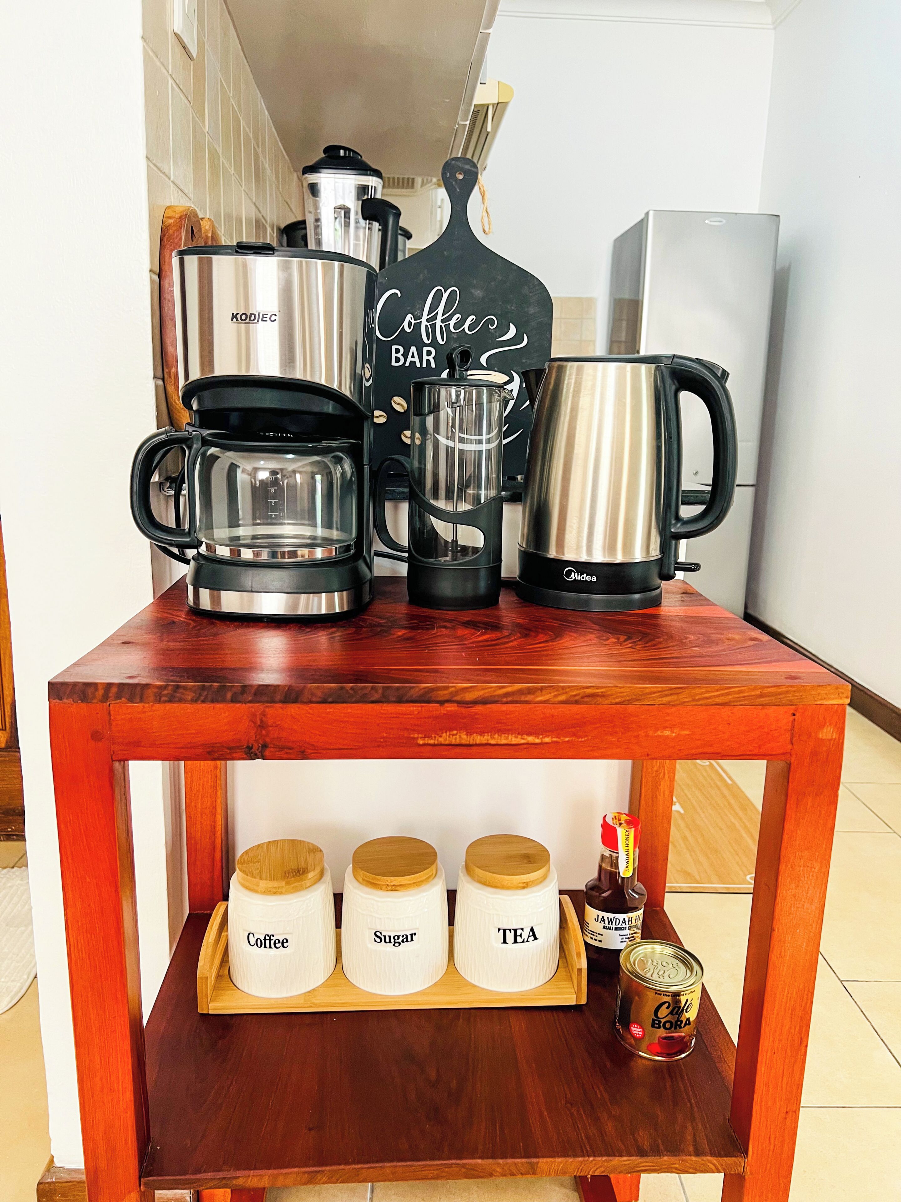 A wooden coffee station with a coffee maker, grinder, and kettle. Below, there are containers labeled coffee, sugar, and tea.