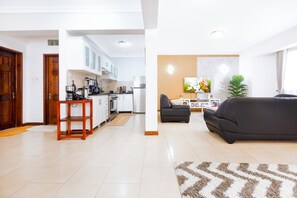 Een moderne keuken met witte kasten, een eethoek met een zwarte bank, en een woonkamer met een groot raam.