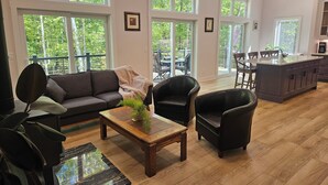 Amplia sala de estar con sofá, dos sillones, una mesa de centro y una cocina con barra y comedor. Grandes ventanales ofrecen vistas al exterior.