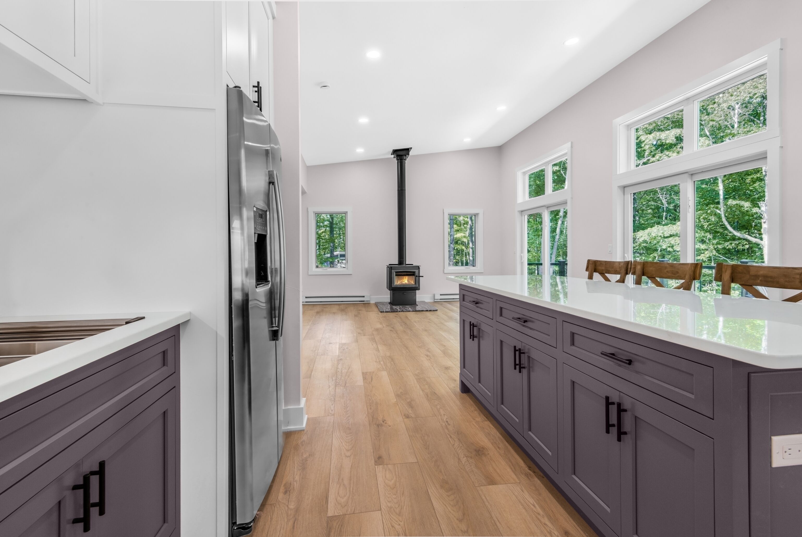 A modern kitchen with a central wood-burning stove, white cabinets, and large windows.