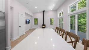 Una habitación amplia con una larga mesa blanca, sillas de madera, chimenea, ventanas grandes y una puerta con un televisor.