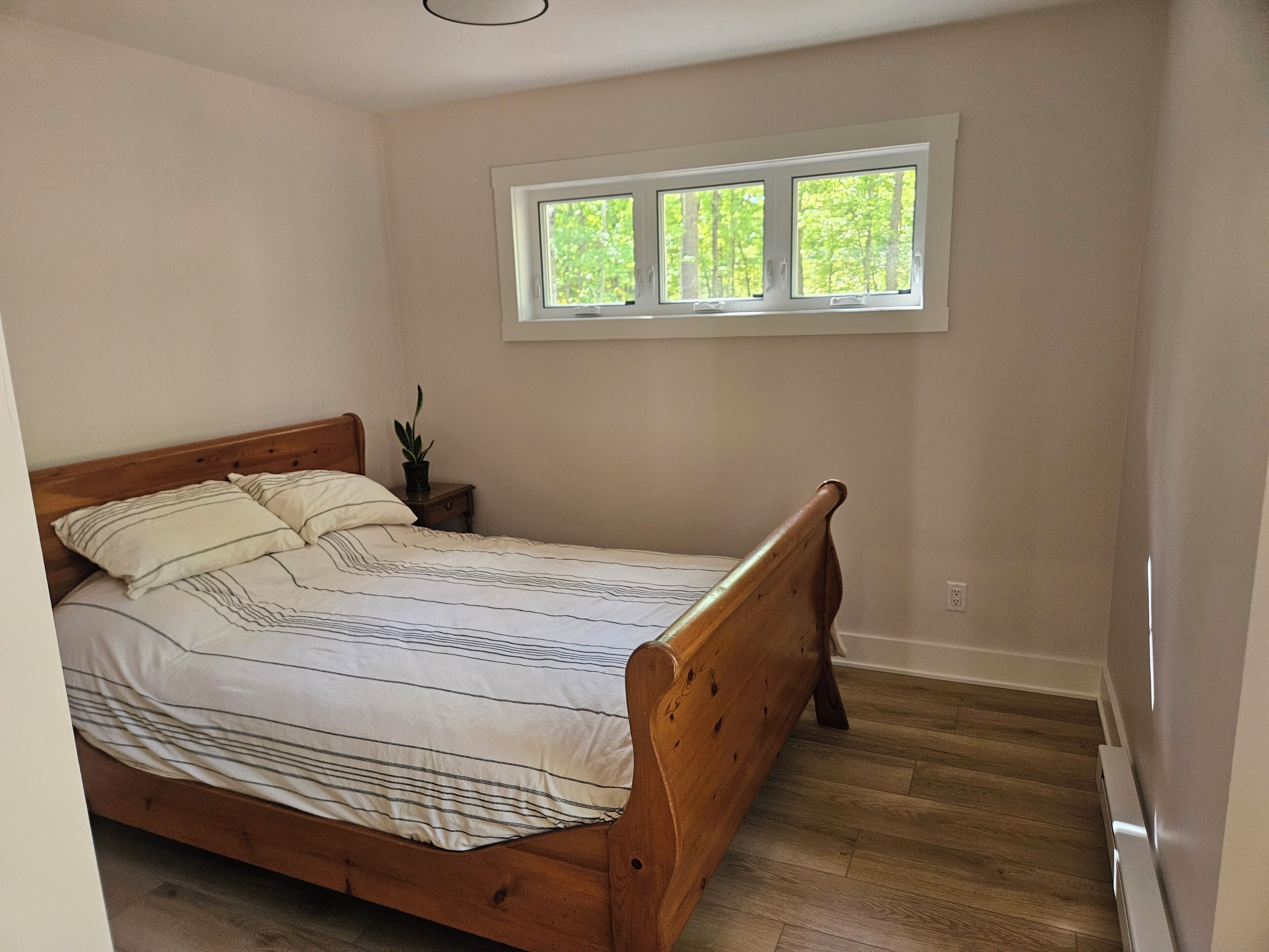 A bedroom with a wooden bed, a nightstand with a plant, and a window with a view of trees.