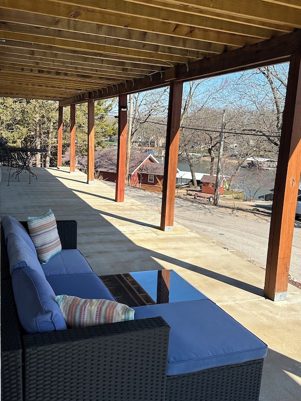 A covered outdoor seating area with a wicker sofa and cushions, wooden pillars, and a view of a residential area with houses and a body of water.