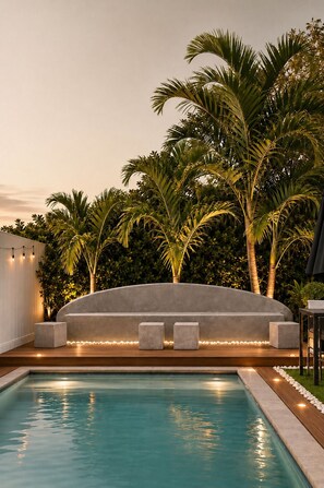 Une piscine dotée d’un espace détente intégré, entourée de palmiers et d’une haie.