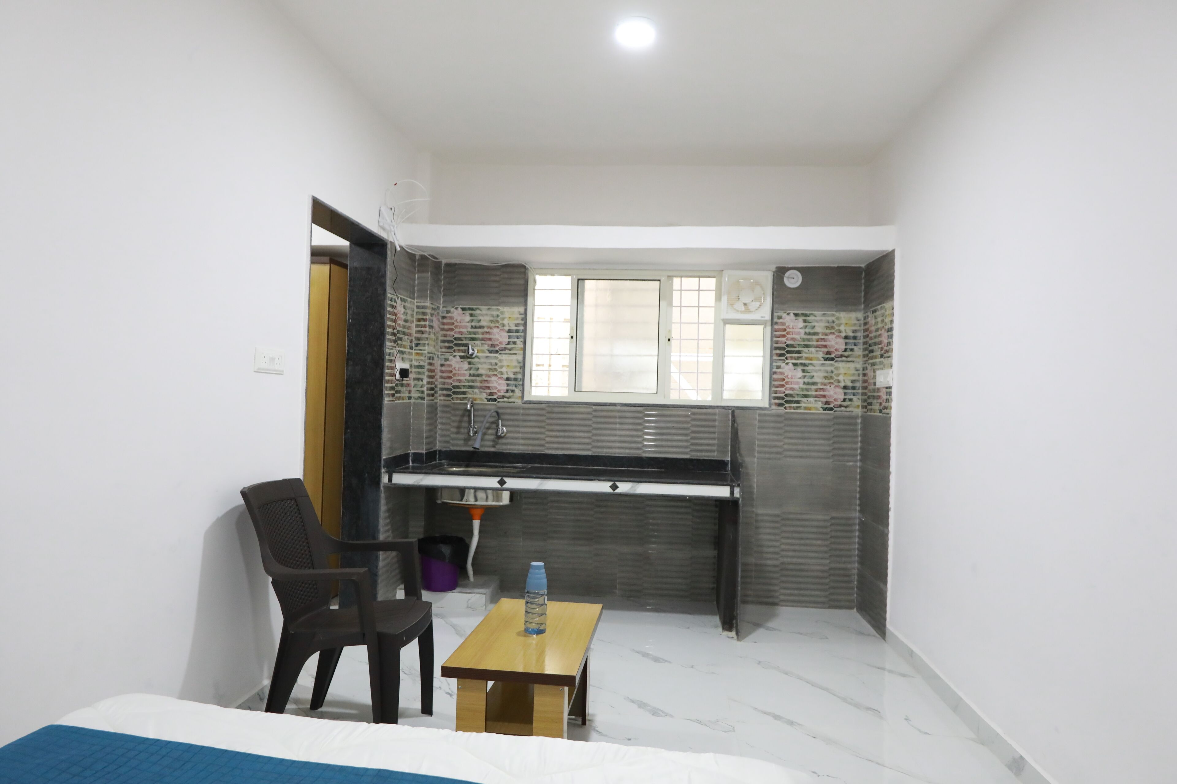 Habitación pequeña con una cama, una mesita con una botella, una silla, un escritorio con un lavabo y una ventana.