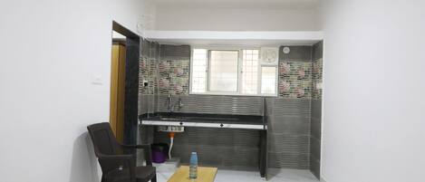 Habitación pequeña con una cama, una mesita con una botella, una silla, un escritorio con un lavabo y una ventana.