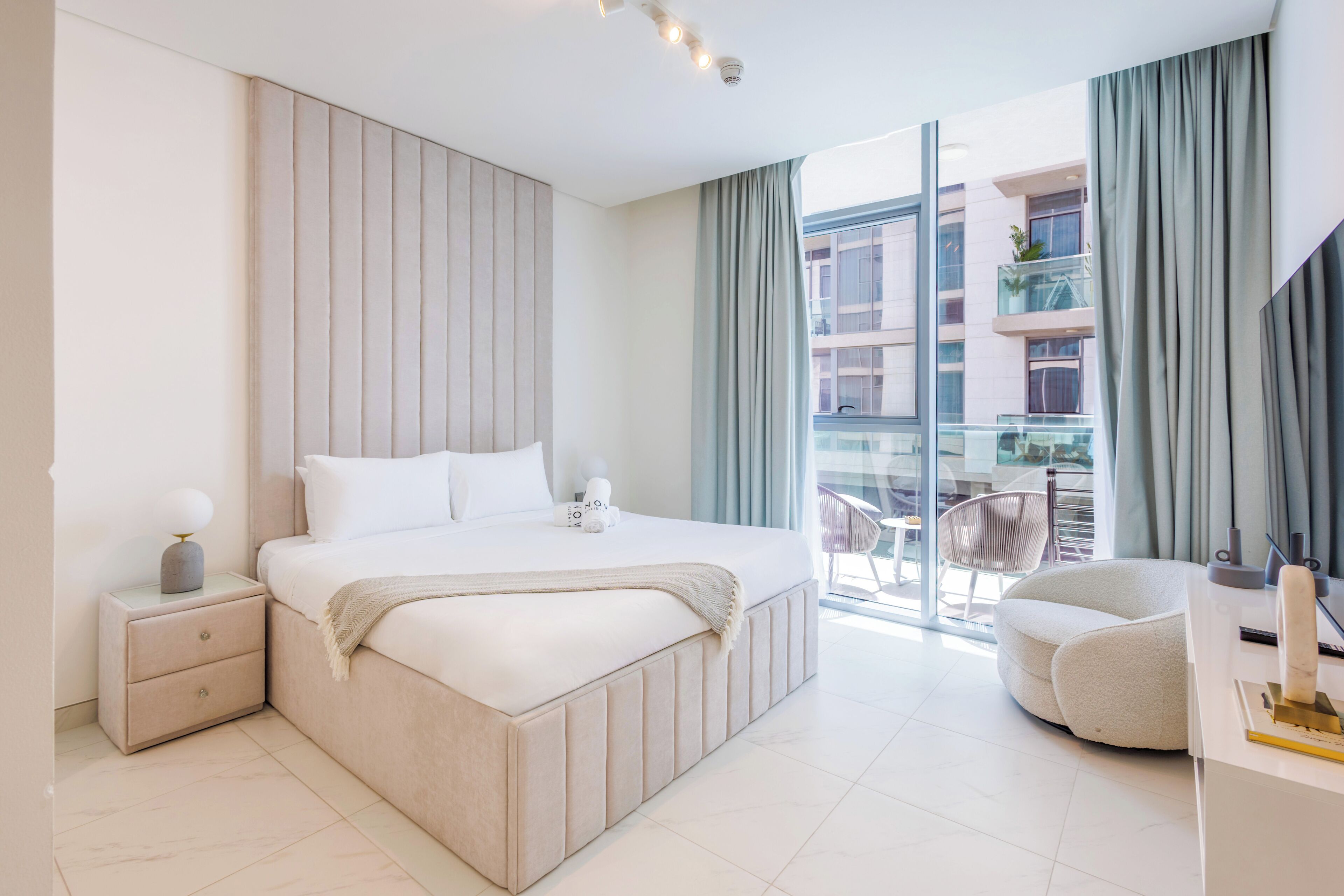 Un dormitorio moderno con una cama grande, una mesita de noche, un escritorio y vistas a la ciudad a través de amplias ventanas.