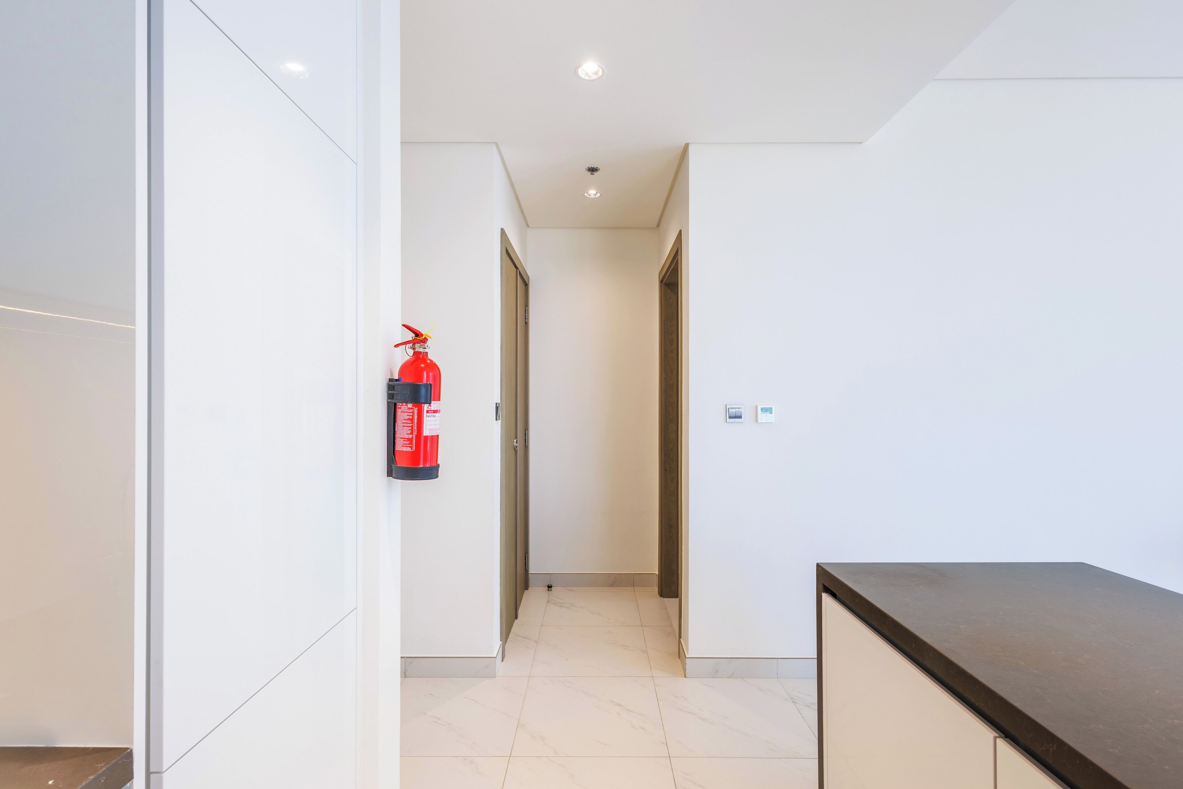 A modern hallway with a fire extinguisher mounted on the wall, a clean tiled floor, and a dark-colored counter on the right.