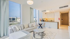 A modern living room with a sofa, a coffee table, and a dining area with chairs. There is a kitchen in the background with a bar counter.