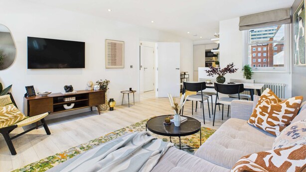 A modern living room with a sofa, coffee table, and a TV mounted on the wall. There is a dining area with chairs and a table visible in the background.