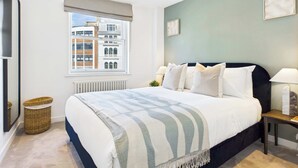 A modern bedroom with a large bed, a view of buildings through the window, and a small bedside table with a lamp. - Chic and cosy apartment near Borough Market (Greater London)