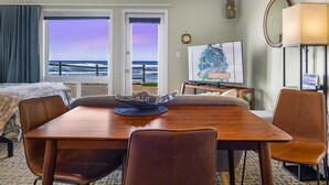 A hotel room with a wooden dining table, two brown leather chairs, a TV on a stand, a lamp, and a view of the ocean through the windows. - Ground Floor Studio, Oceanfront Patio, Kitchenette (Lincoln City)
