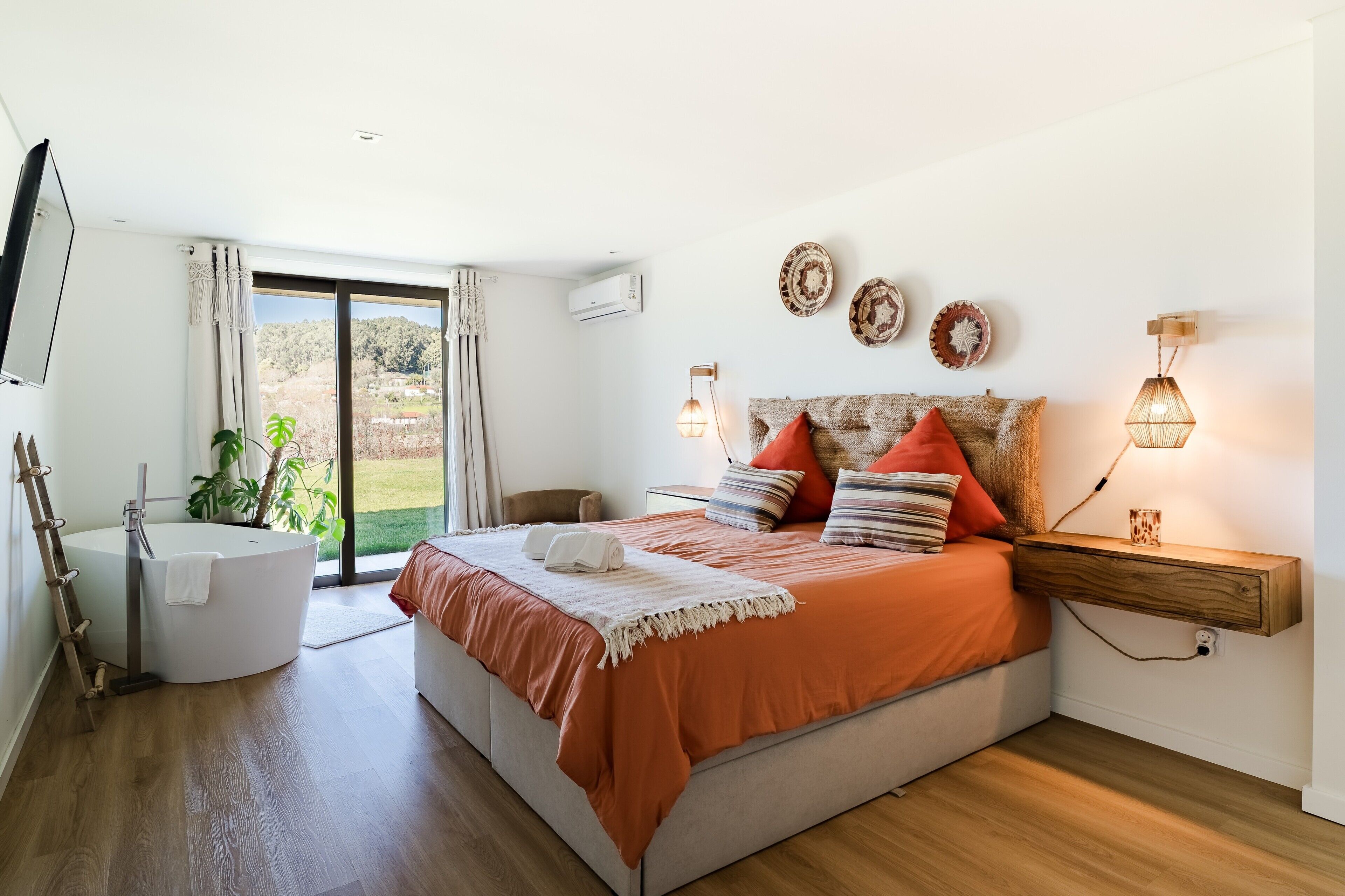 A bedroom with a bed, a built-in bathtub, a wooden floor, a large window, and a view of the outdoors.