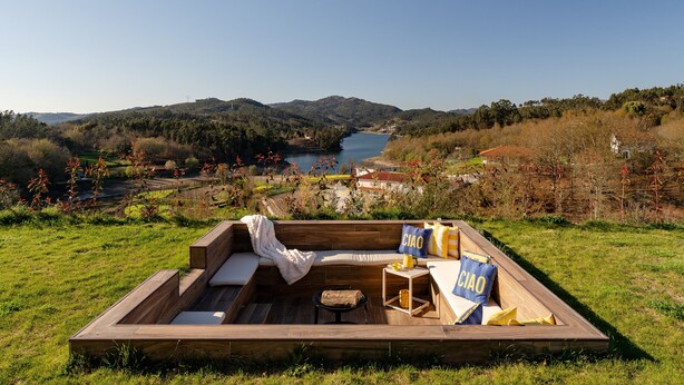 Terraza de madera con sofá, cojines y una mesita, con vistas a un lago y colinas.