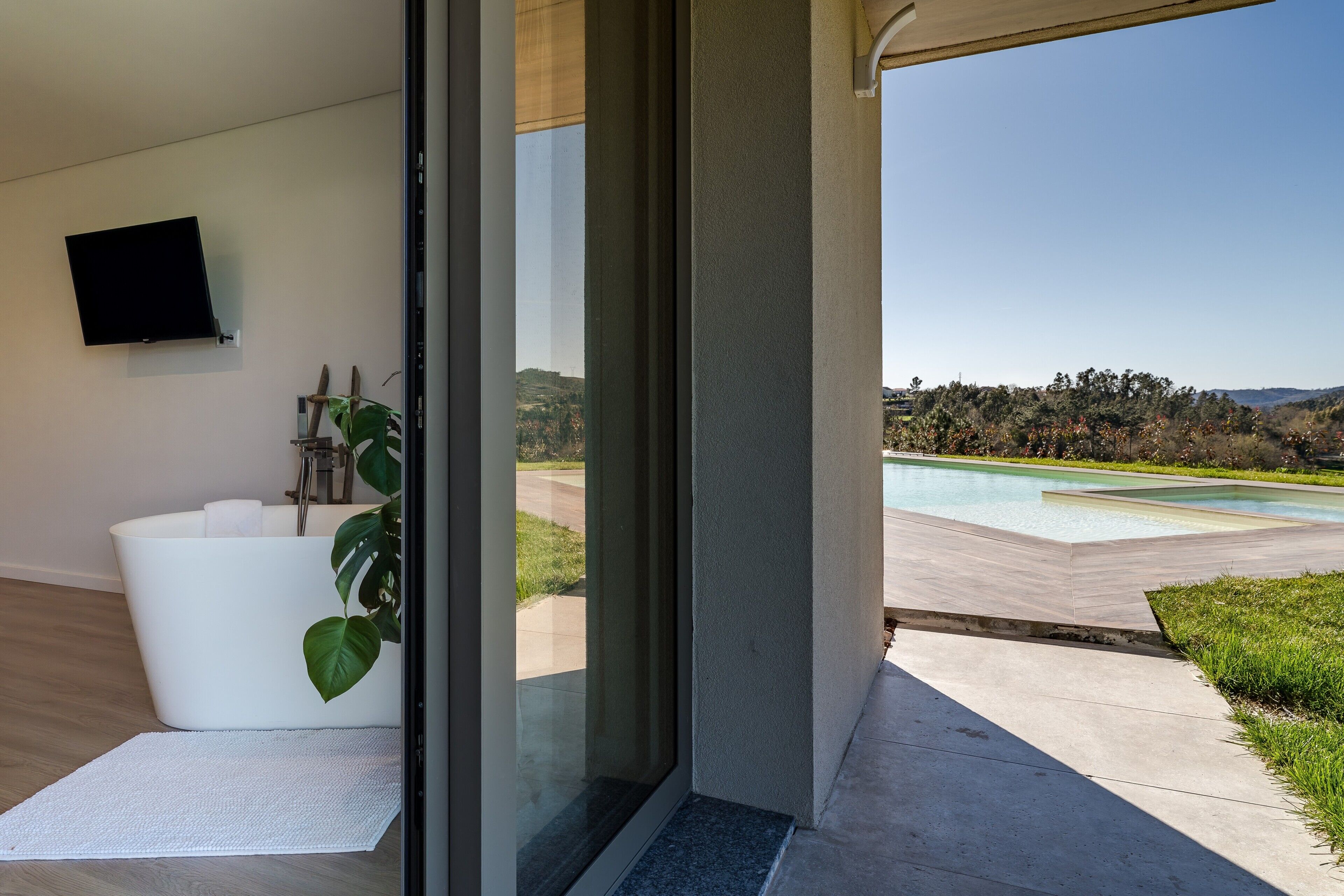 Un bagno moderno con una vasca, una TV a schermo piatto e vista su una piscina e delle colline.
