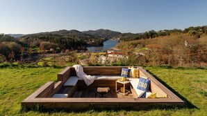 A wooden deck with a sofa, cushions, and a small table, overlooking a lake and hills.