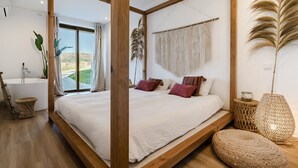 A bedroom with a large bed, a view of the outdoors, a bathtub, and a macrame wall hanging.