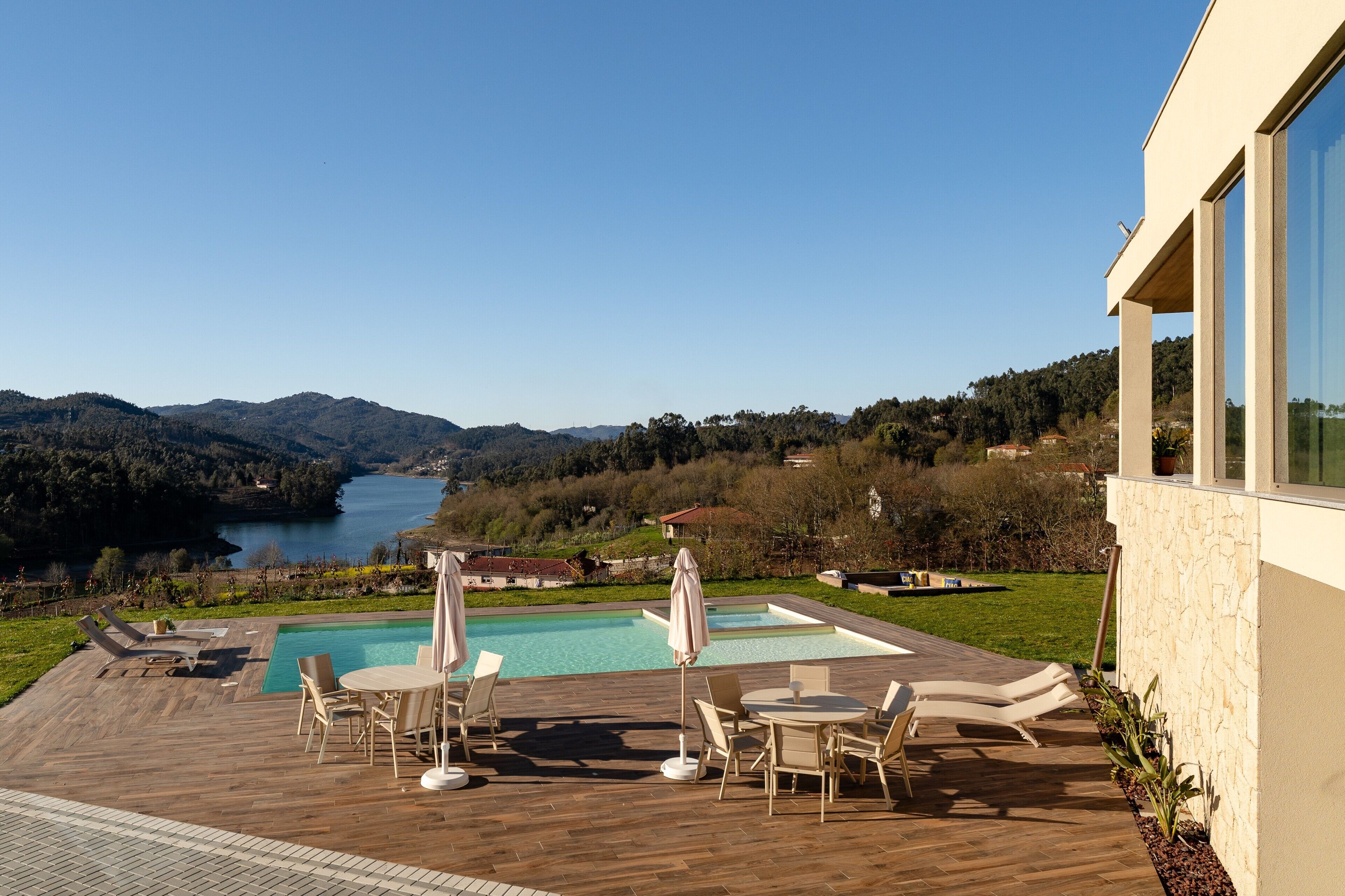 Un área de piscina con sillones de descanso y sombrillas, una terraza de madera y vistas a un lago y montañas.