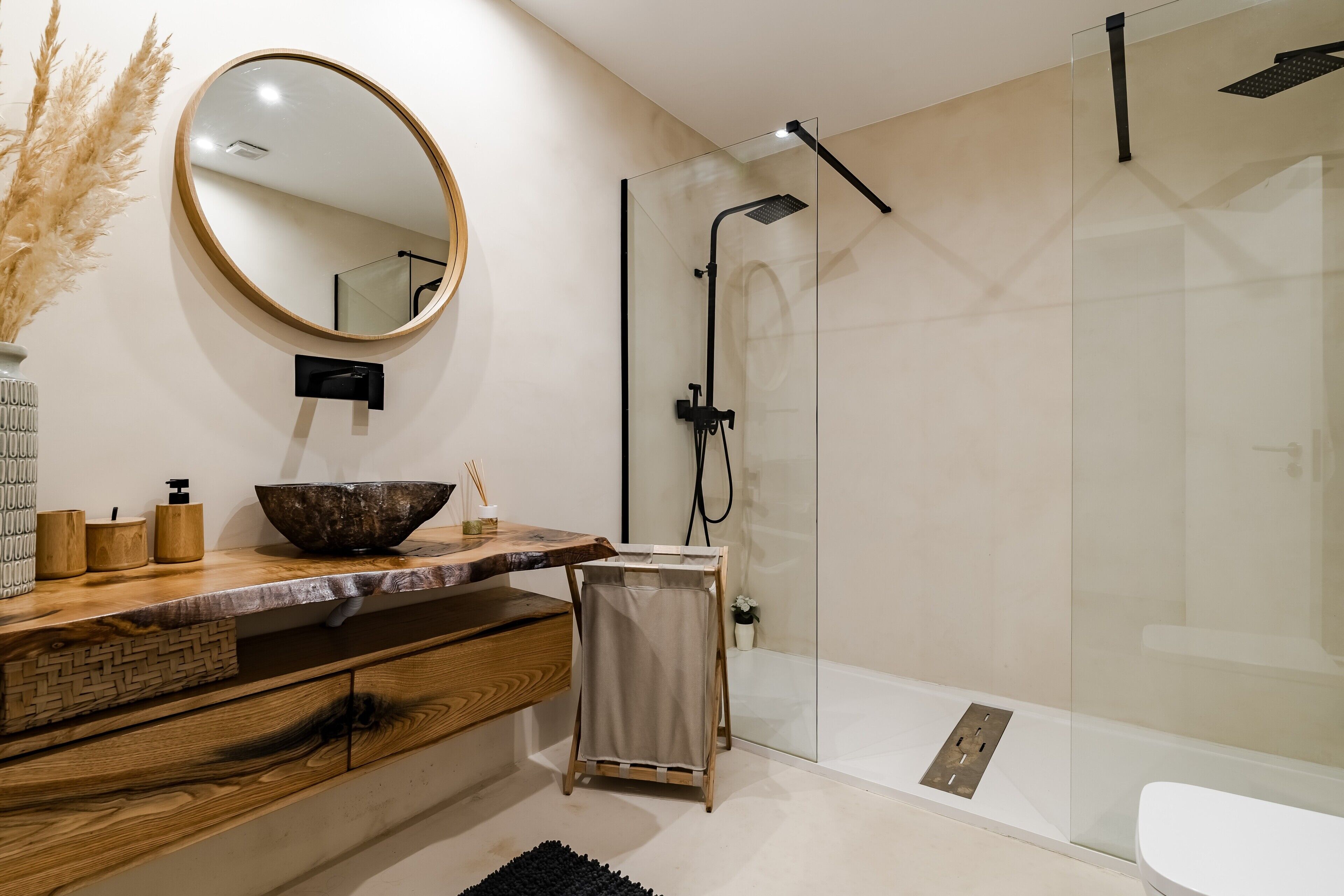 Un baño moderno con tocador de madera, espejo redondo, cabina de ducha con mampara de vidrio y un inodoro blanco.