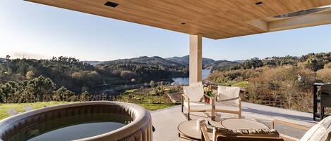 A modern outdoor patio with a hot tub, seating, and a scenic view of hills and a river.
