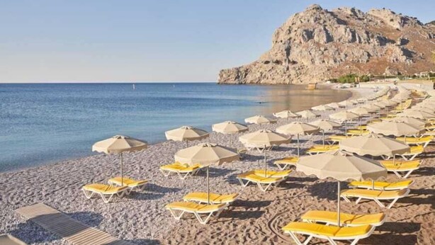 Une plage bordée de rangées de transats et de parasols jaunes, une falaise rocheuse en arrière-plan et une mer bleue et limpide.