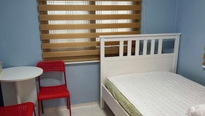 Een slaapkamer met een enkel bed, een klein tafeltje, twee rode stoelen en een raam met jaloezieën.