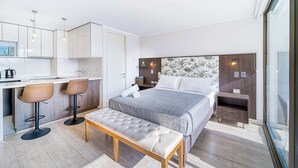 A modern hotel room with a bed, a bench, a desk with two bar stools, a small table, and a large window.
