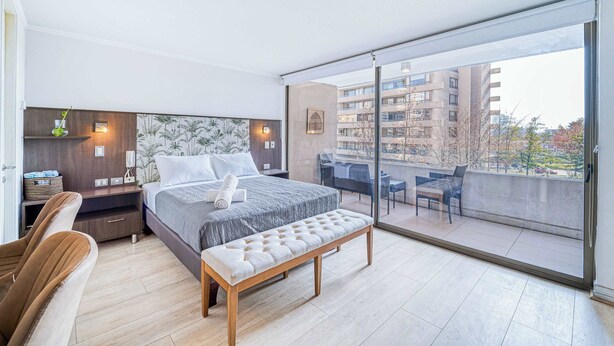 A modern bedroom with a large bed, a bench, a desk, and a balcony with a view of buildings.