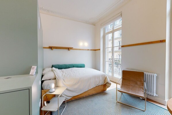 Une chambre d’hôtel comprenant un lit, une table de chevet, une lampe, une chaise, un radiateur et une fenêtre donnant sur des immeubles.