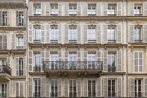 Un immeuble à plusieurs étages, doté de volets uniformes, de balcons ornés de garde-corps en fer forgé et de détails architecturaux classiques.