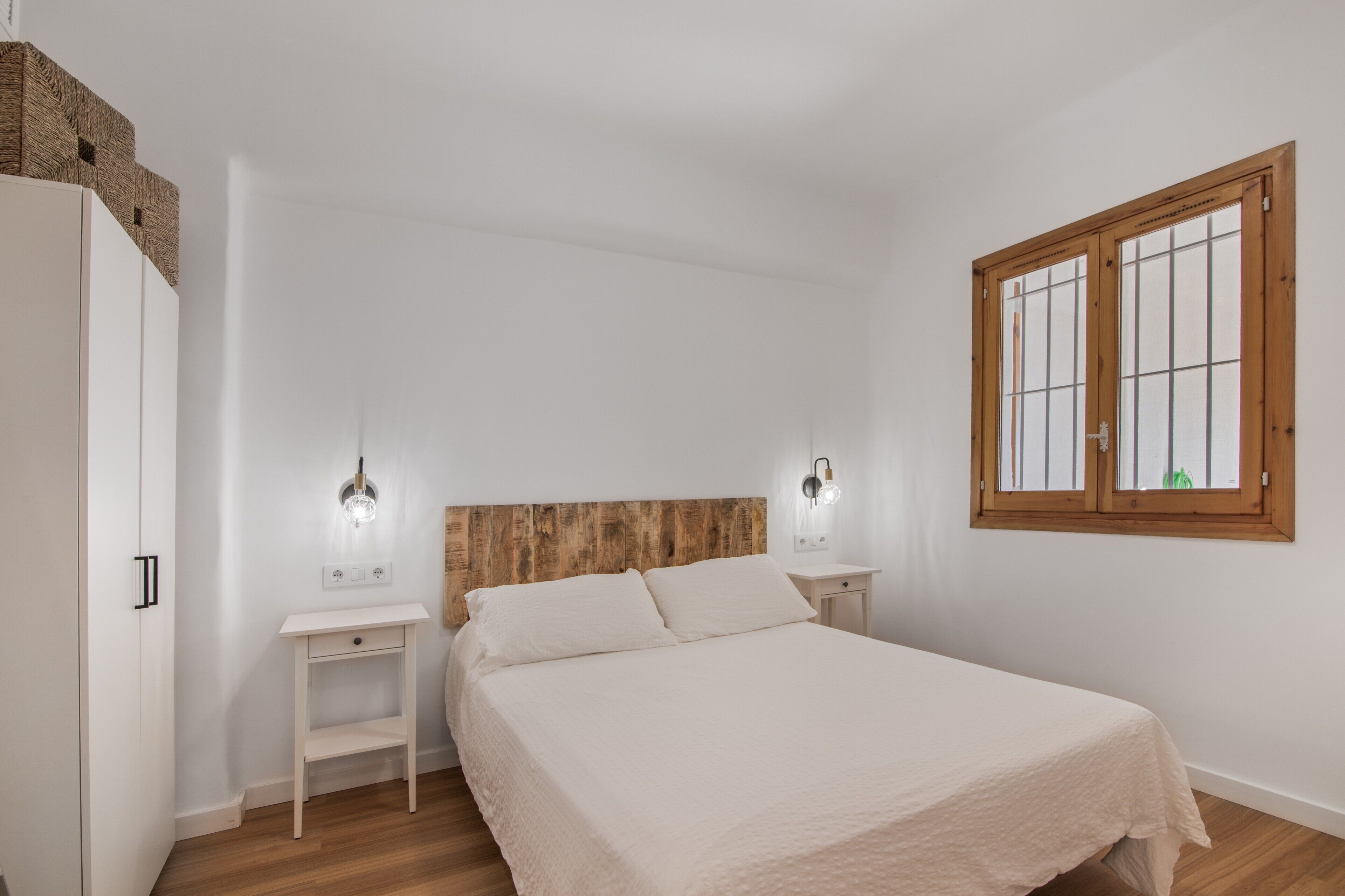 Een slaapkamer met een houten hoofdbord, een bed met een witte dekbedovertrek, twee nachtkastjes met wandlampen, een raam met houten kozijnen en een witte kledingkast.