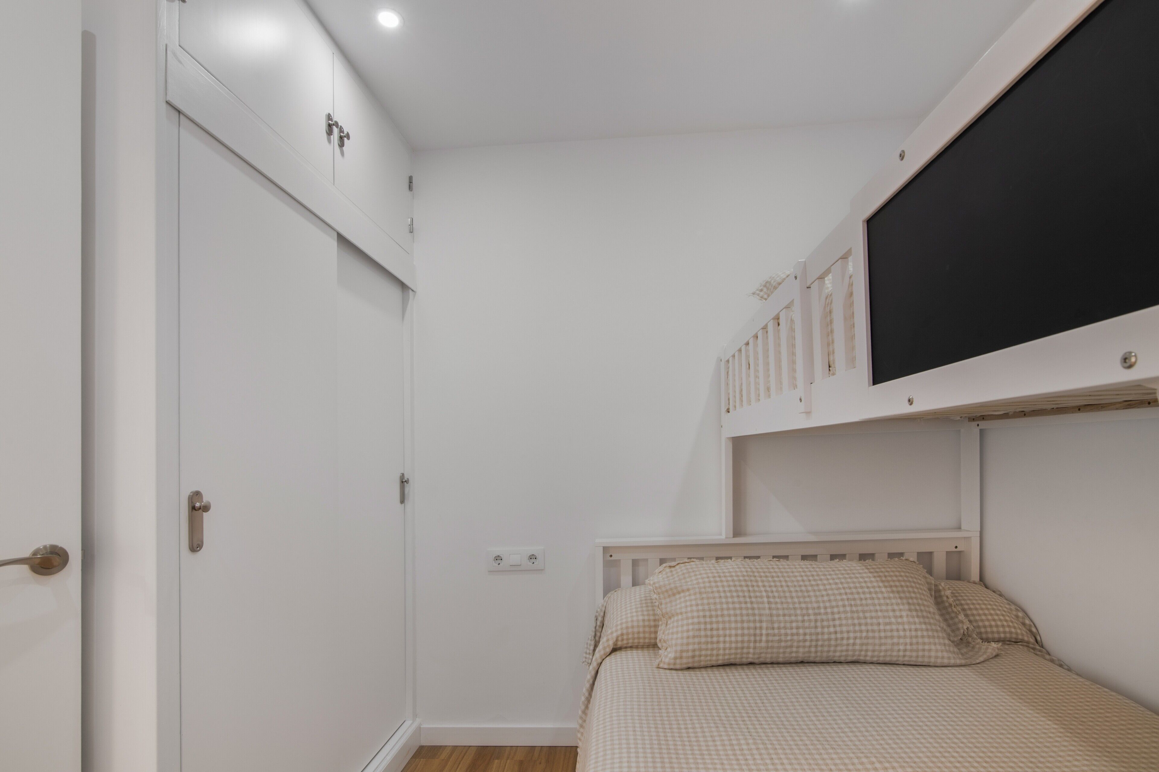 A bedroom with a single bed, a white wardrobe, a mounted television, and a door with a handle.