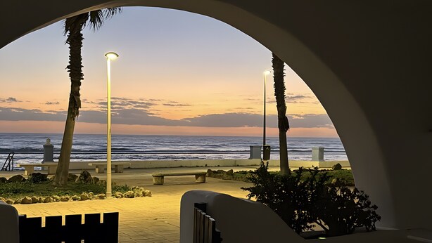 A view through an archway to a beach with a sunset, palm trees, and a bench. - Holiday Home 'Primera Linea En Matalascañas' with Sea View, Wi-Fi and Air Conditioning (Almonte)