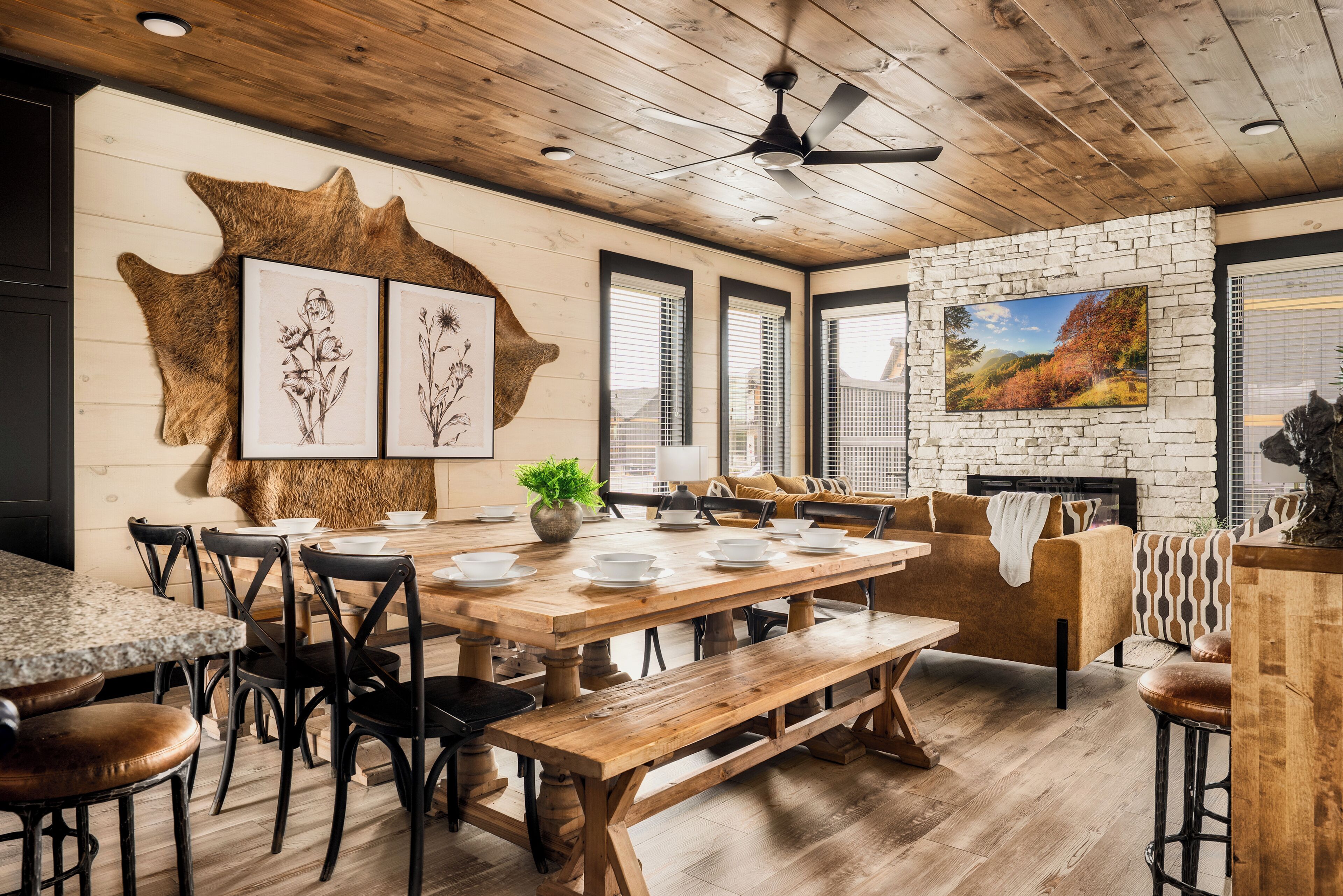 Un coin repas rustique avec une table en bois, des bancs et des chaises noires. Les murs sont ornés d’illustrations botaniques encadrées, et on y trouve un ventilateur de plafond ainsi qu’une grande fenêtre munie de stores.
