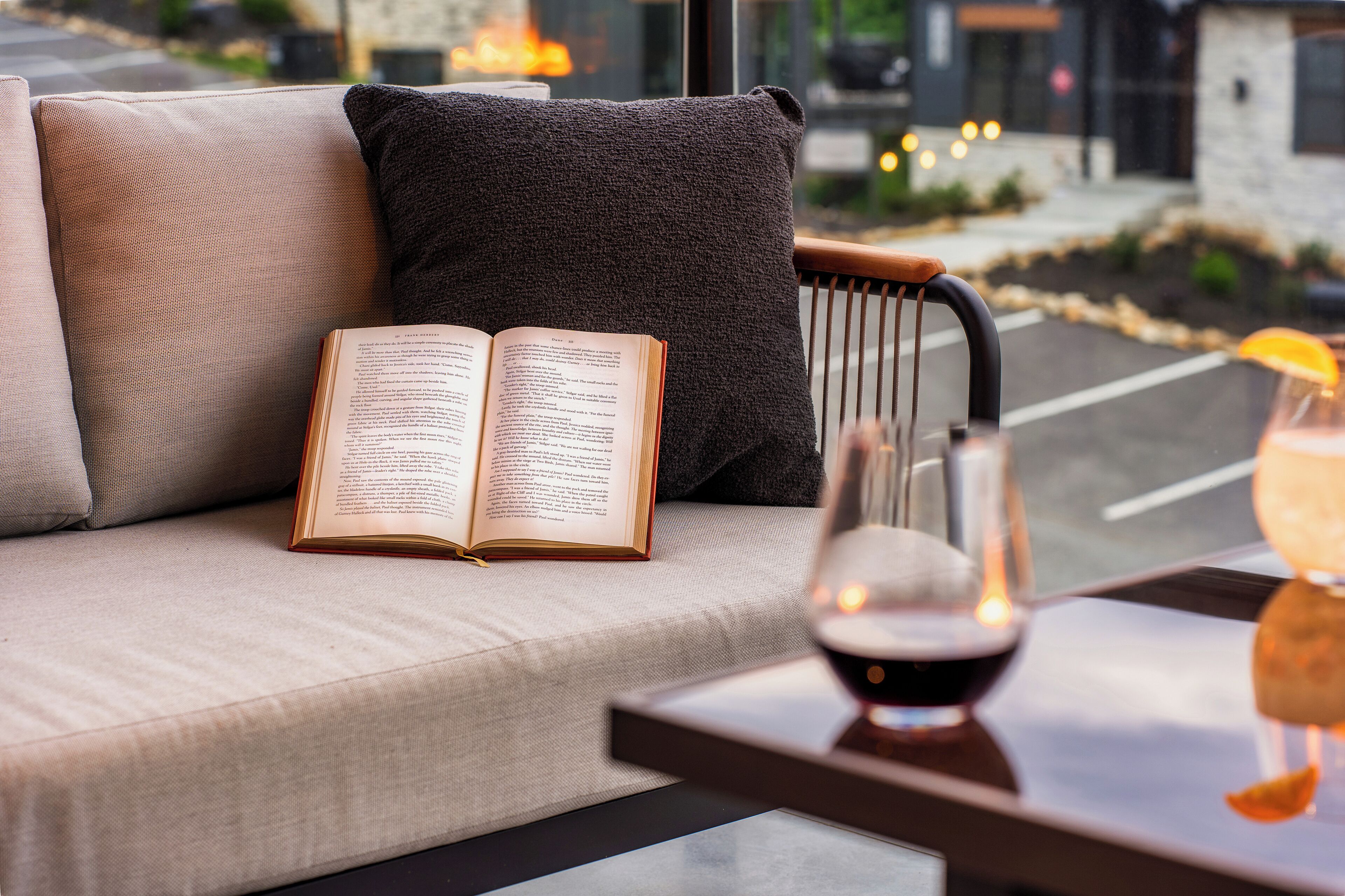 An open book on a sofa with a cushion, a glass of drink on a table, and a view of the outdoors.